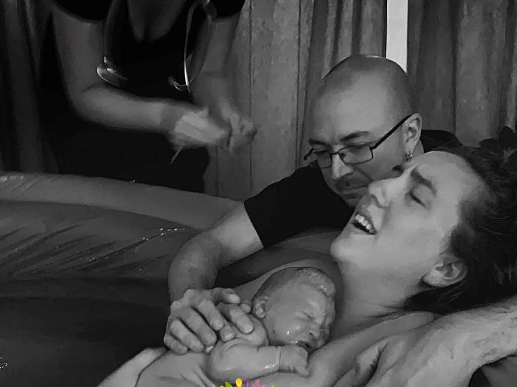 Hypno-mom Dana in birth tub just after birth holding her newborn baby with her partner kneeling behind her.
