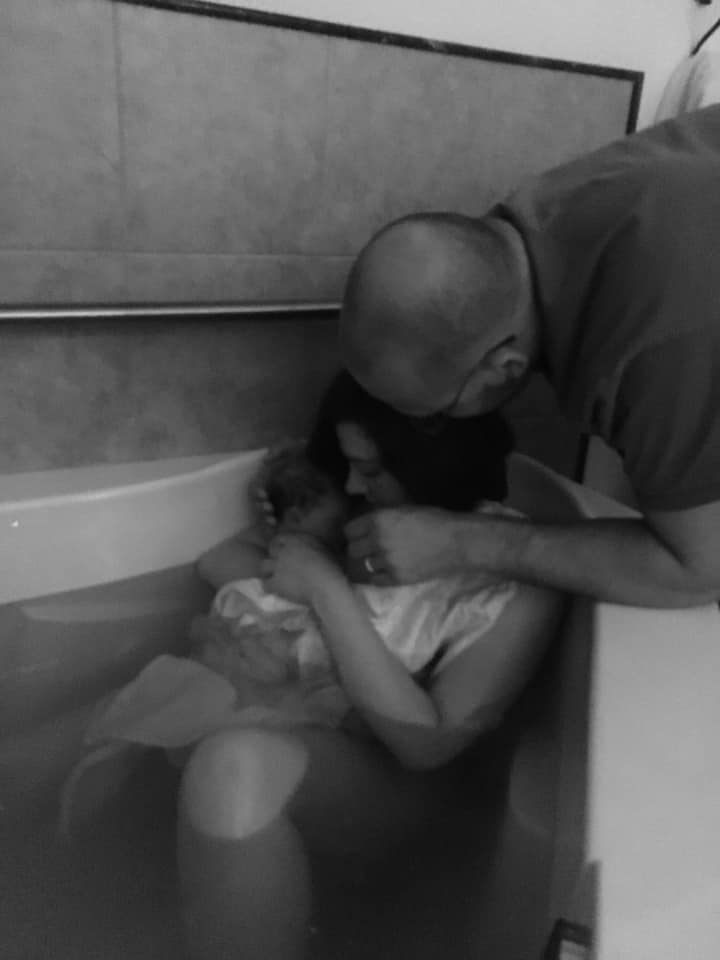 Hypno-mom Francesca in birth tub with newborn baby and partner standing behind her leaning over.