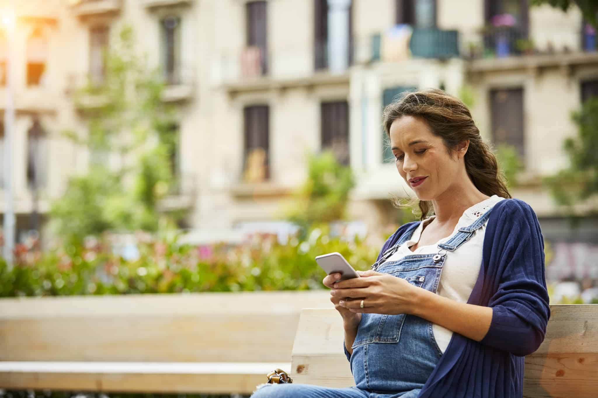 Pregnant woman wearing overalls, sitting on a bench and looking at her phone.