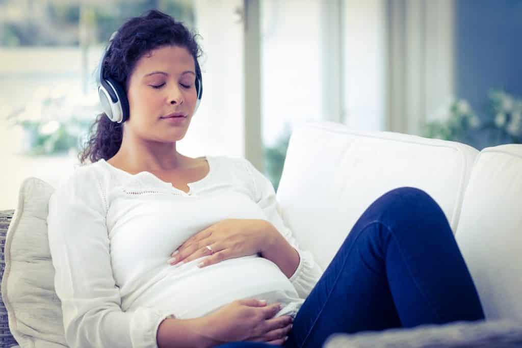 Pregnant person with her eyes closed in a white shirt and jeans laying back on a sofa with headphones on and her hands on her belly.