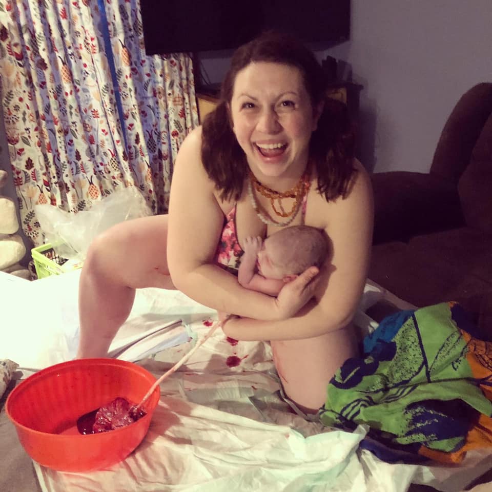 Hypno-mom kneeling holding newborn just after birth with huge smile and her placenta in a bowl in front of her.