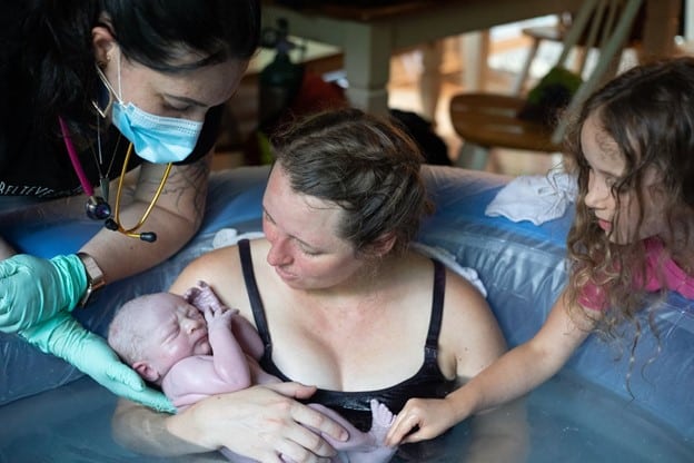 New parent just after giving birth in tub holding newborn with older child and midwife helping