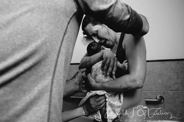 Black and white photo immediately after a waterbirth kneeling in tub holding baby while midwife and birth partner assist