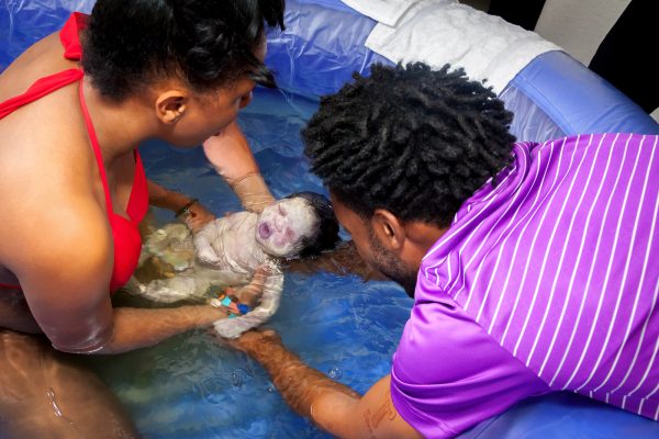New parents welcoming baby in pool of water at home during a homebirth with help from their midwife.