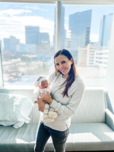 new parent holding new baby in front of large window overlooking a city