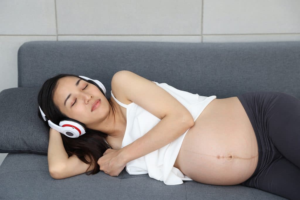 young mom, pregnant woman lying down on sofa with headphones listening to hypnosis